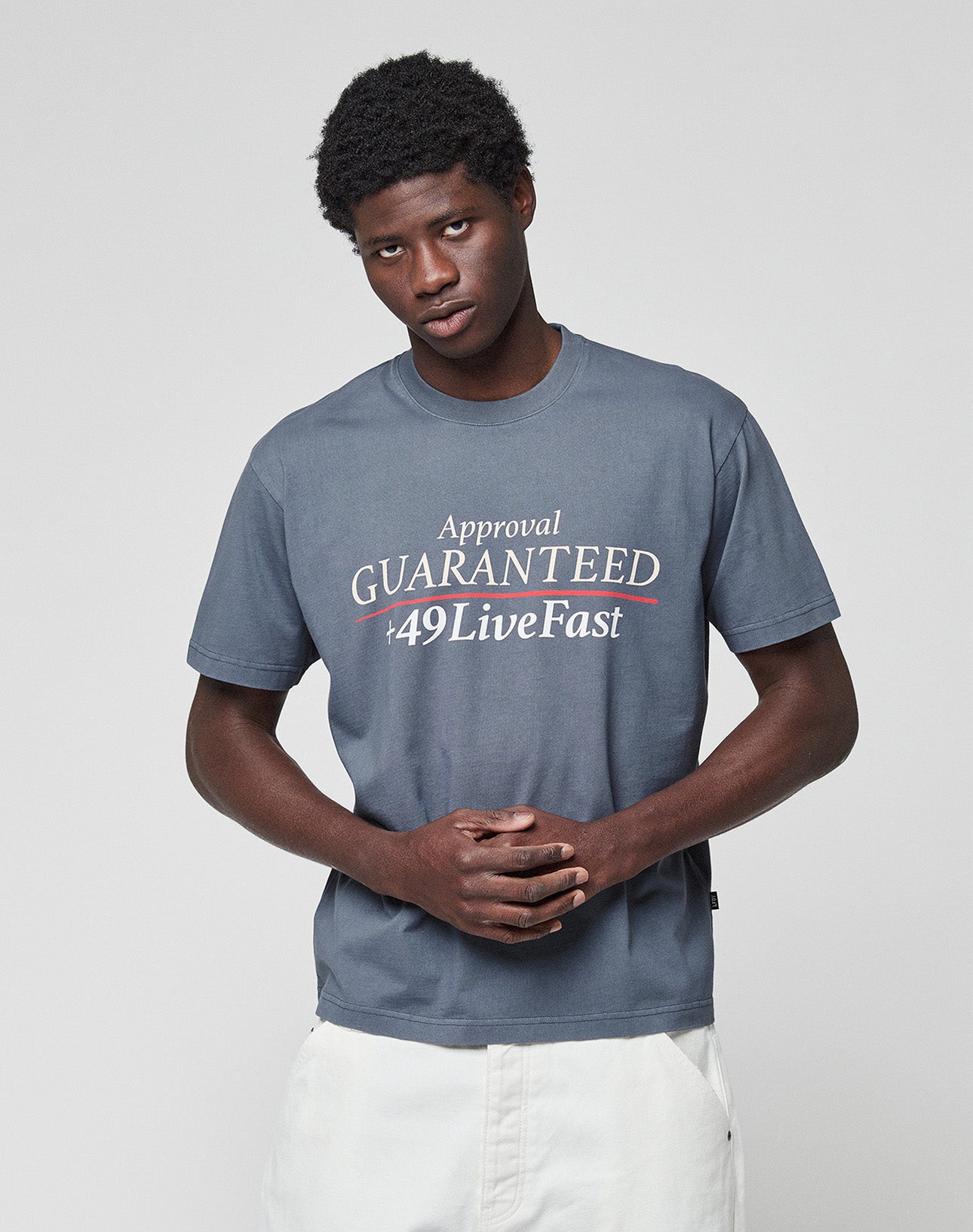 A young man with short curly hair wears the LIVE FAST DIE YOUNG LFDY Guaranteed Tee—gray, 100% Baumwolle, bold “Approval GUARANTEED +49LiveFast” front print—and white shorts, standing against a light gray background with clasped hands.