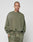 A person with a shaved head and face tattoos wears the LIVE FAST DIE YOUNG Basic Chest Sweater in oversized olive green with matching sweatpants featuring a handcrafted fade effect, standing against a plain light gray background.