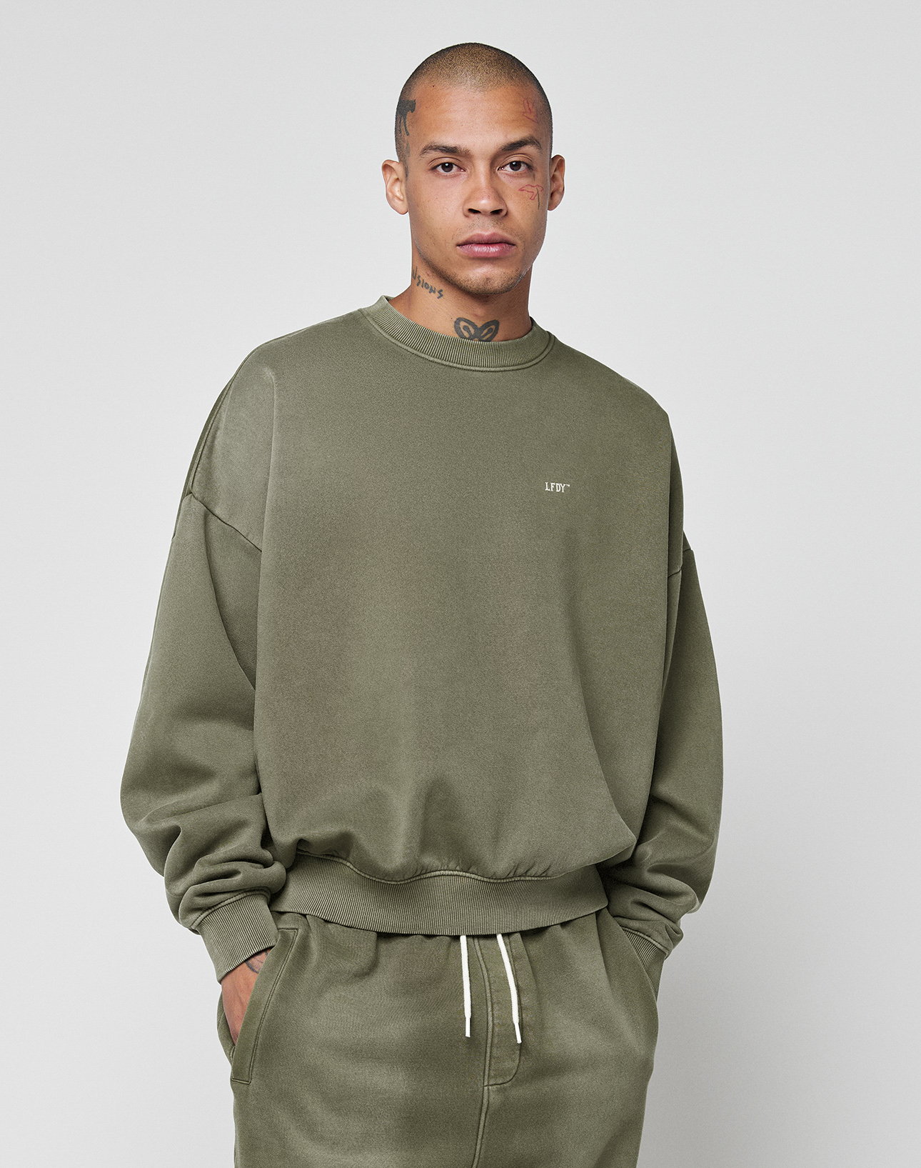 A person with short hair and visible face and neck tattoos wears the LIVE FAST DIE YOUNG Basic Chest Sweater in oversized olive green with a unique handgefertigter Fade-Effekt, standing against a plain light background.