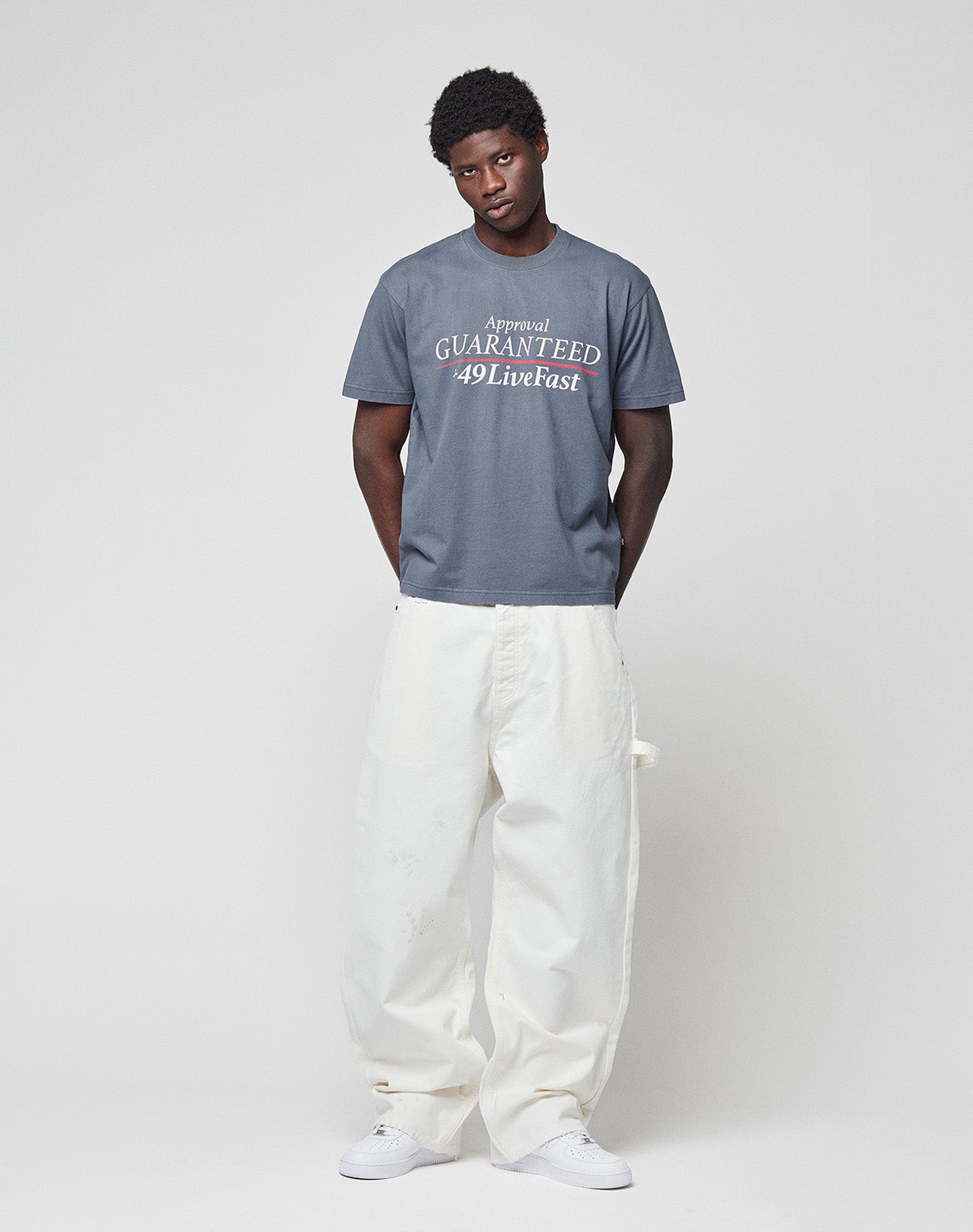 A person with short curly hair stands against a plain light background, wearing LIVE FAST DIE YOUNG Worker Pants in white 100% Baumwolle with a Carpenter Pocket and a gray T-shirt with white text, paired with white sneakers.
