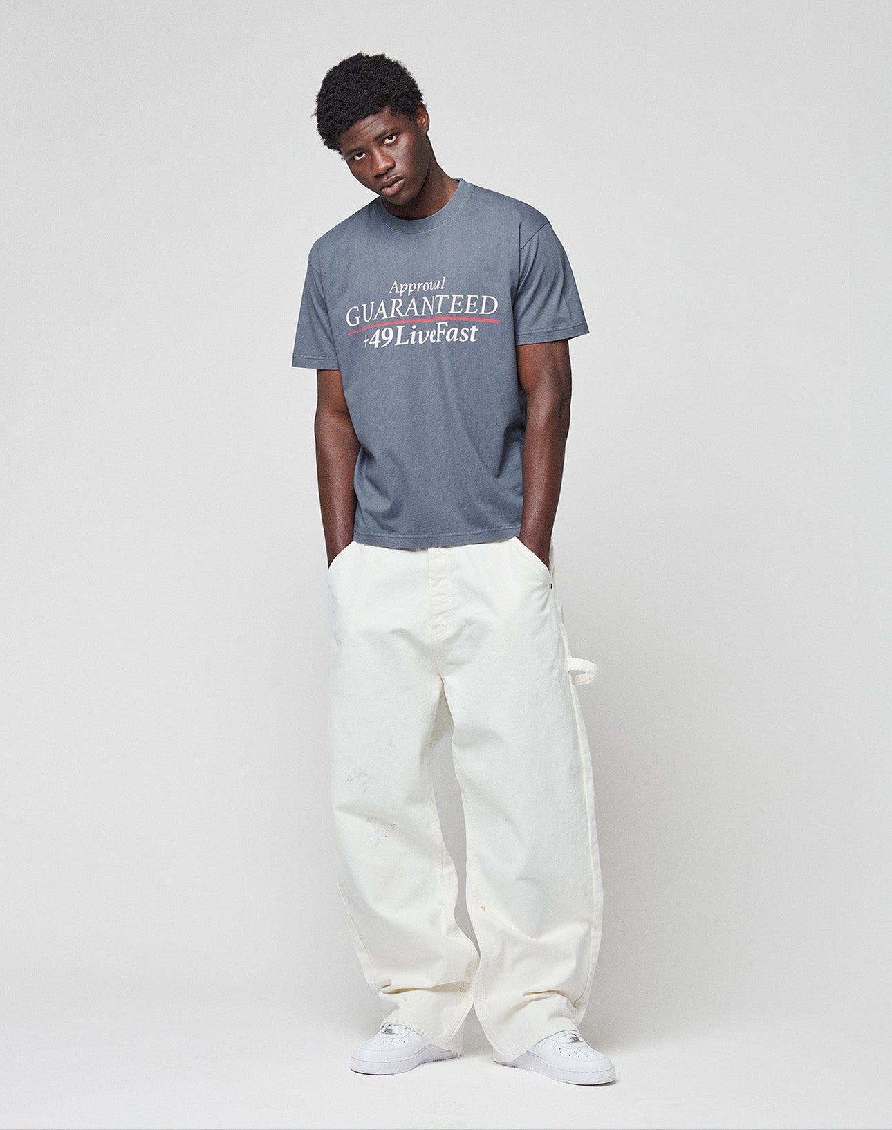 A person stands against a plain light background, wearing the LIVE FAST DIE YOUNG LFDY Guaranteed Tee in regular fit with “Approval GUARANTEED 49 Livfast” frontprint, paired with loose white pants and white sneakers. The tee is 100% cotton.
