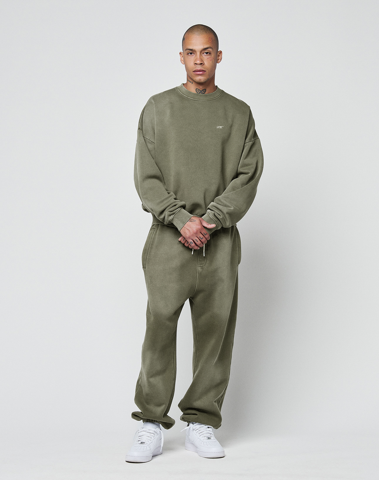A person stands against a light background, wearing the LIVE FAST DIE YOUNG Basic Chest Sweater in olive green and matching sweatpants with white sneakers. Their relaxed pose highlights the oversize fit and Faded Look as they face the camera.