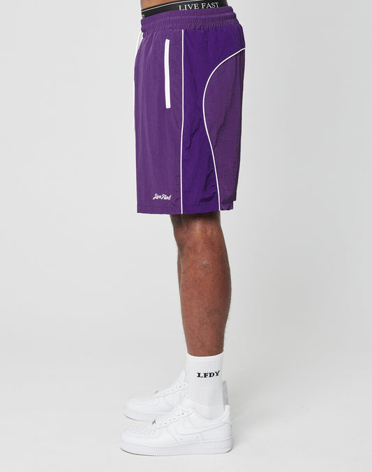 A person in LIVE FAST DIE YOUNG Beach Shorts with white piping, an elastic waistband, white LFDY socks, and white sneakers stands sideways against a plain white background.