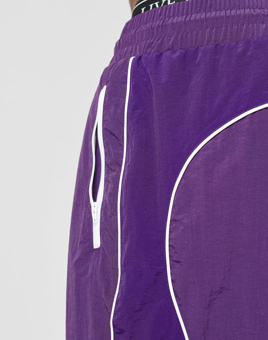 Close-up of a person wearing LIVE FAST DIE YOUNG Beach Shorts in purple nylon with white piping, side zipper pocket, black Elastik-Bund waistband with white branding, and a crinkled texture for a regular fit.