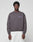 A man stands facing forward against a light gray background, wearing the LIVE FAST DIE YOUNG LFDY Network Sweater in an oversized dark gray fit with a small purple chest graphic, paired with white pants.