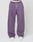 A person stands wearing LIVE FAST DIE YOUNG’s Basic Wide Jogger in purple, featuring wide fit, white drawstrings, an elastic waistband with "LIVE FAST" text, and white sneakers. Background is plain and light-colored.
