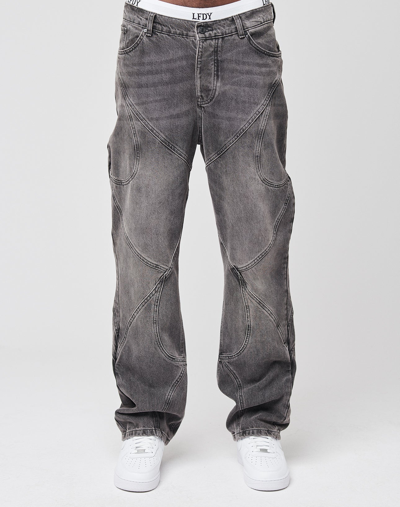 A person wears LIVE FAST DIE YOUNG Tribal Wide Denim—loose, dark gray jeans with tribal panels and curved seams—paired with white sneakers and visible white underwear, standing out against a plain, light background.