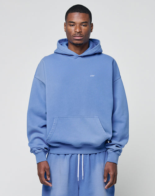 A man faces forward in a matching light blue LIVE FAST DIE YOUNG Basic Evidence Hooded sweatshirt and sweatpants with an oversize fit, front pocket, and small chest text, set against a plain background to highlight the relaxed style.