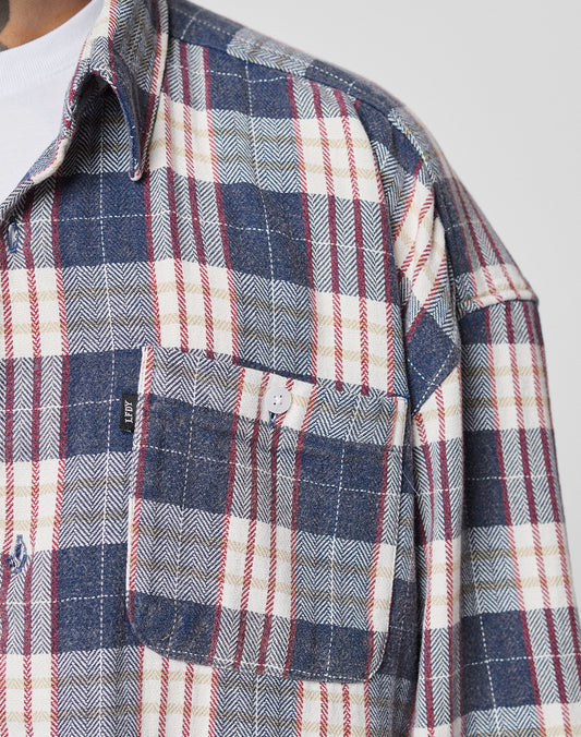 Close-up of someone wearing the LIVE FAST DIE YOUNG Vintage Flannel, an oversized blue, white, and red plaid shirt with a front button pocket, small black tag, zweiteiliger Hemdkragen, styled over a white T-shirt.