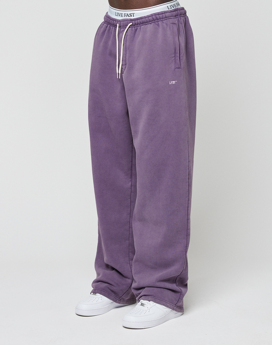 A person wears LIVE FAST DIE YOUNG Basic Wide Joggers in loose purple with a white drawstring and "LIVE FAST" elastic waistband, paired with white sneakers against a plain white background.