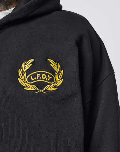 Someone wearing the LFDY Team Hooded Zip by LIVE FAST DIE YOUNG, an oversize-fit black hoodie with a yellow laurel wreath logo, multilogo Stickereien, and L.F.D.Y embroidered on the chest.