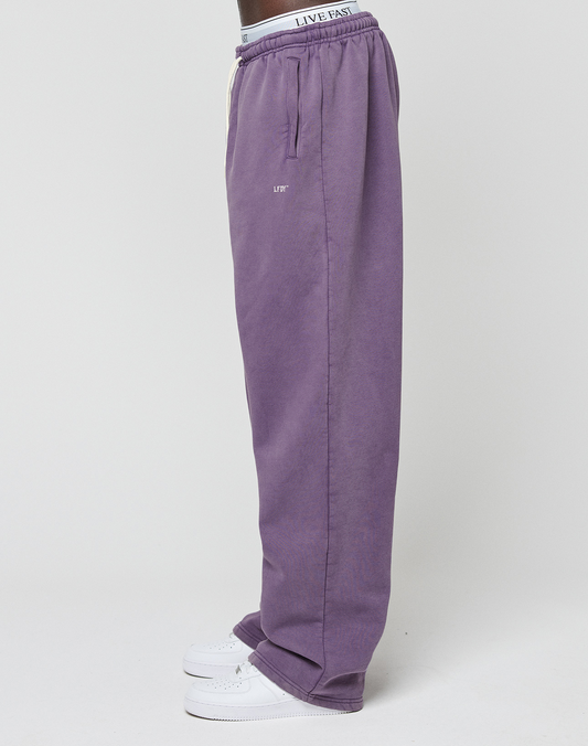 A person stands sideways against a plain light background wearing LIVE FAST DIE YOUNG Basic Wide Jogger in purple, white sneakers, and underwear featuring a visible elastikbund waistband with the text LIVE EAST.