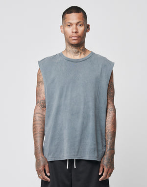A man with short hair and tattoos wears the LIVE FAST DIE YOUNG Basic Muscle Shirt, a loose-fit, sleeveless gray top in 100% Baumwolle, and black shorts. He stands against a plain light gray background, facing forward with a neutral expression.