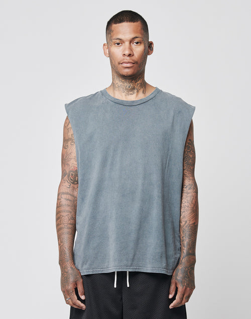 A man with short hair and tattoos wears the LIVE FAST DIE YOUNG Basic Muscle Shirt, a loose-fit, sleeveless gray top in 100% Baumwolle, and black shorts. He stands against a plain light gray background, facing forward with a neutral expression.