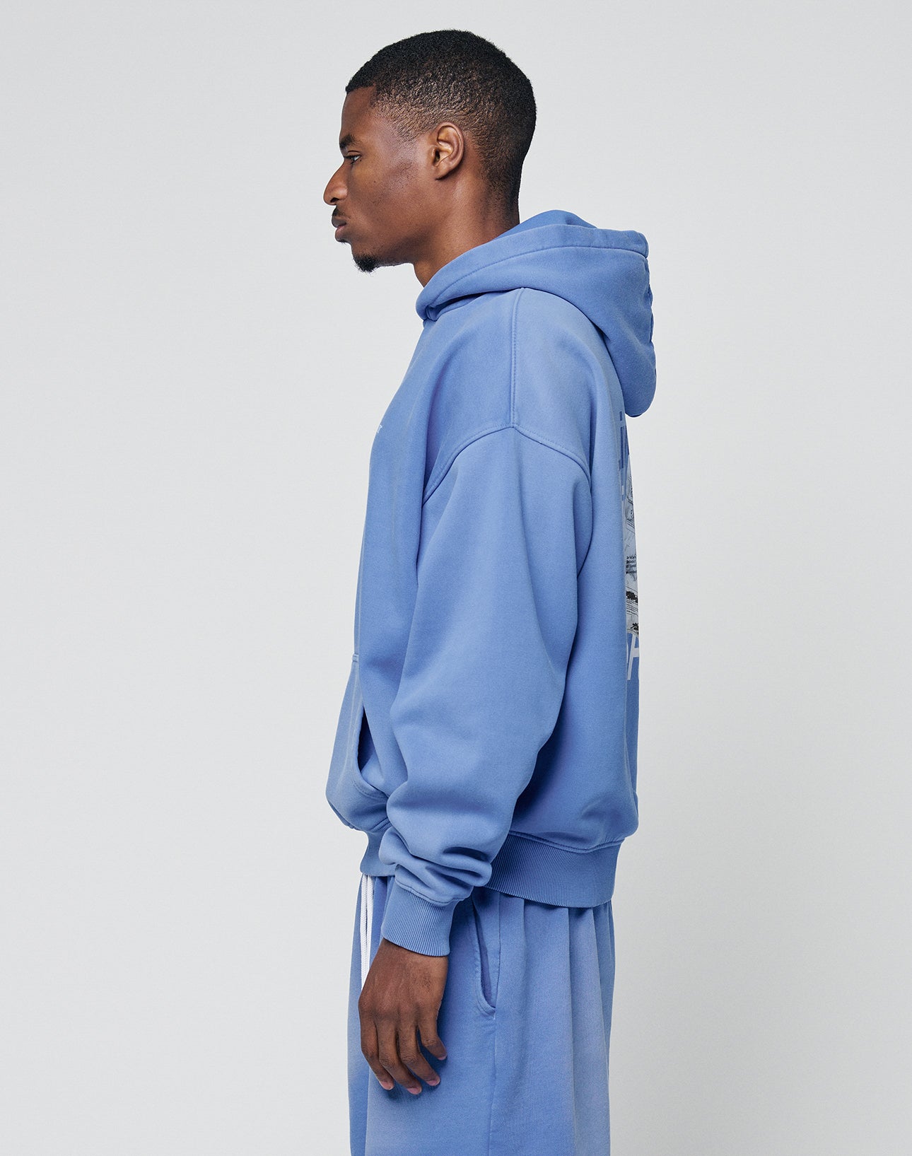 A young man stands in profile wearing the LIVE FAST DIE YOUNG Basic Evidence Hooded in an oversize fit with matching blue sweatpants, both featuring a subtle faded look, set against a plain light gray background.