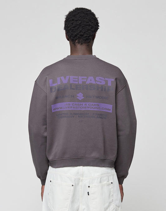 A person faces away, wearing the LFDY Network Sweater by LIVE FAST DIE YOUNG—an oversize dark gray cotton sweatshirt with purple "LIVEFAST DEALERSHIP" front and back prints—paired with white pants, set against a plain light background.