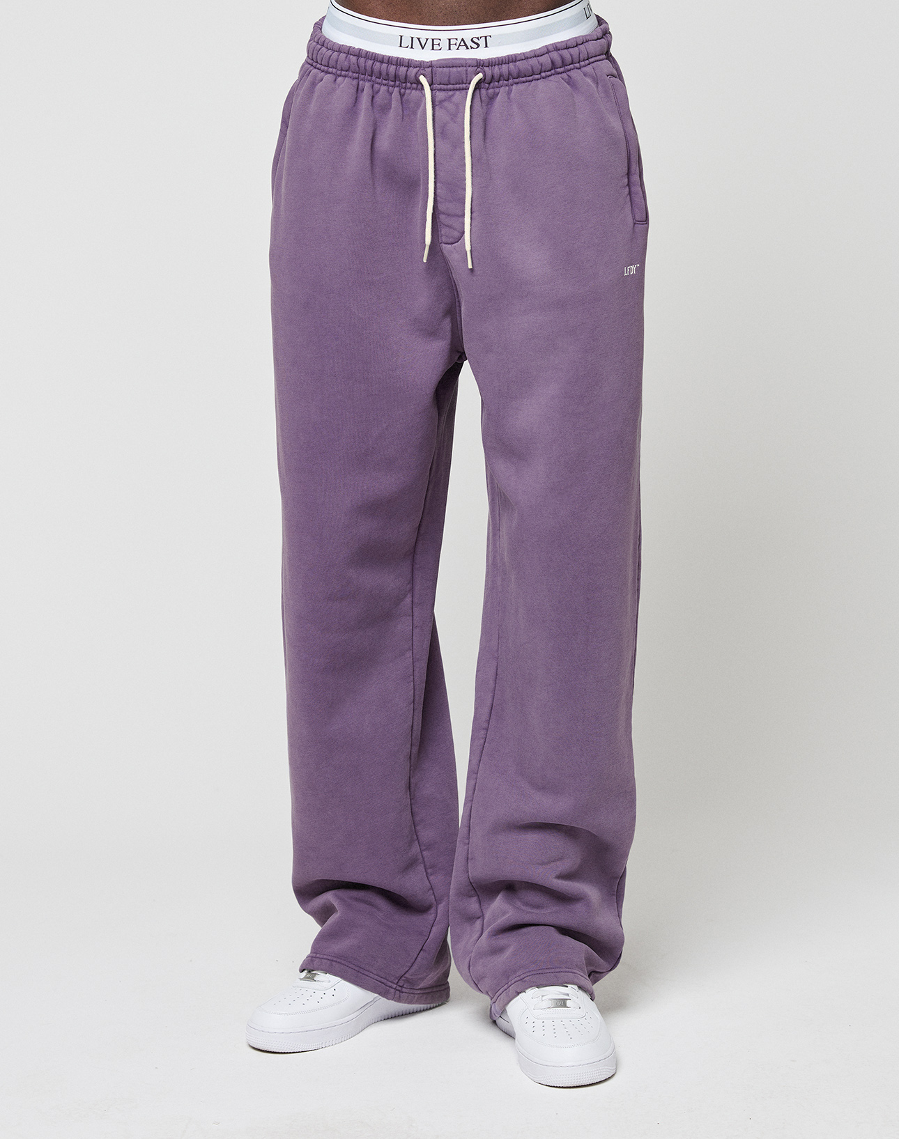 A person wearing the LIVE FAST DIE YOUNG Basic Wide Jogger in purple with a wide fit, elastic waistband, white drawstrings, white sneakers, and a visible LIVE FAST logo-print waistband stands against a plain white background.