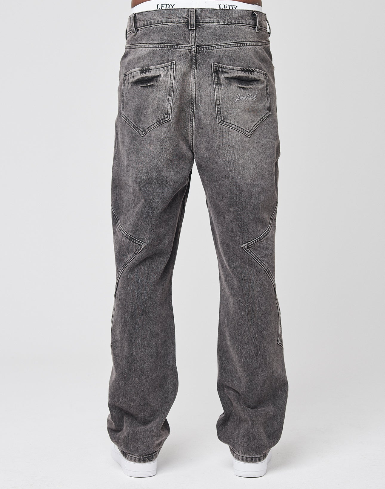 A person is seen from the back wearing LIVE FAST DIE YOUNG Tribal Wide Denim—loose-fitting, faded black jeans in 100% Baumwolle with worn details, visible-stitched back pockets, and styled with white sneakers.