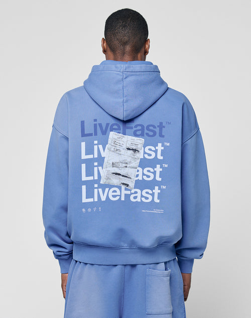 A person wearing the LIVE FAST DIE YOUNG Basic Evidence Hooded, an oversized blue hoodie with a faded finish and repeated LiveFast prints in white and navy, plus overlapping receipts and symbols on the back, faces away from the camera.