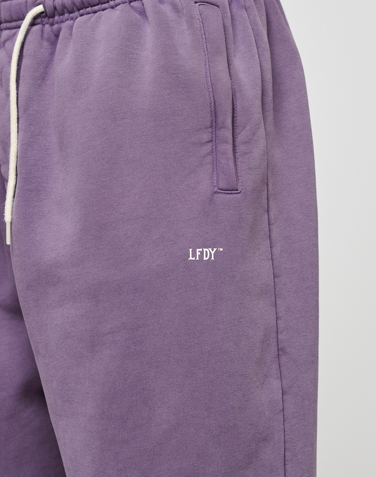 Close-up of a person wearing the LIVE FAST DIE YOUNG Basic Wide Jogger in purple, featuring a white drawstring and embroidered LFDY logo near the pocket.