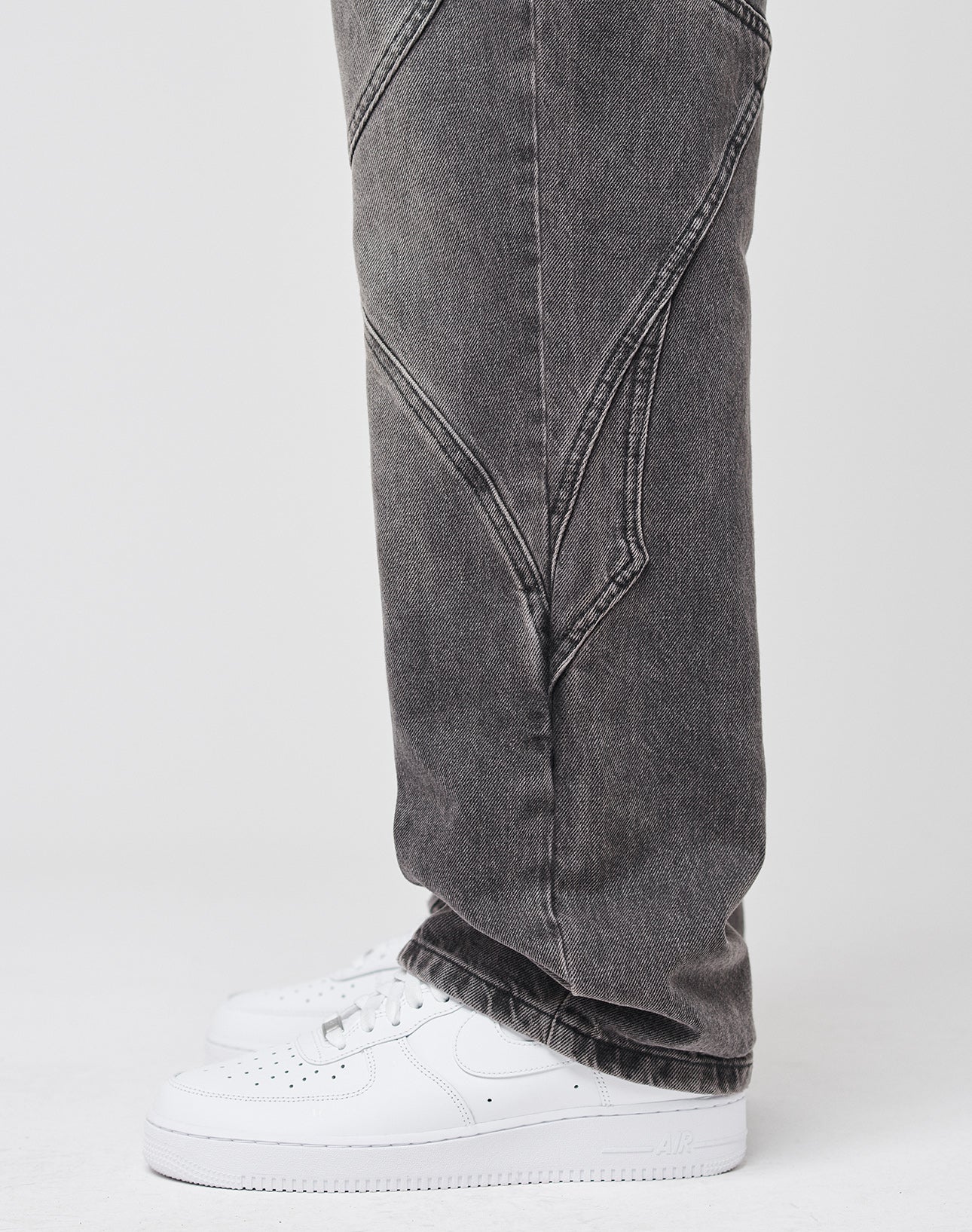 A person wears LIVE FAST DIE YOUNG’s Tribal Wide Denim—faded black, baggy jeans with diagonal seams and tribal panels—paired with white sneakers against a plain light background.