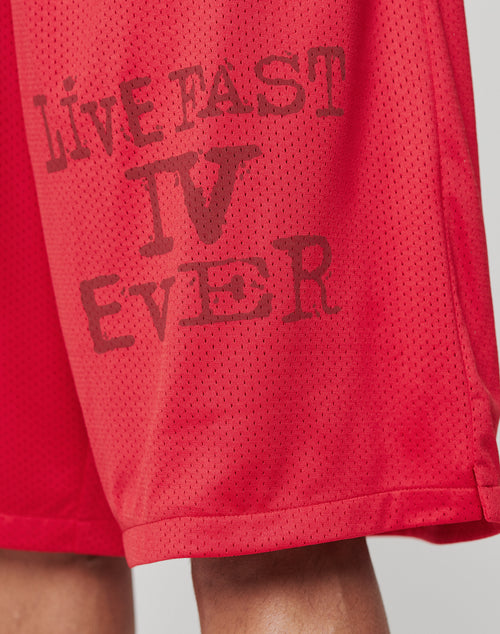 Close-up of LIVE FAST DIE YOUNG’s Live Fast IV Ever Shorts in red athletic mesh, oversized fit, elastic waistband, and LIVE FAST IV EVER faded black print. Polyester. Part of a person’s leg is visible at the bottom.