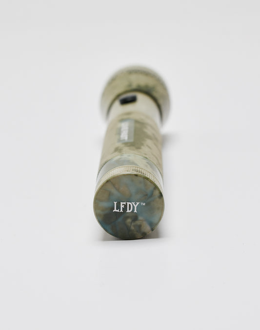 A camo-patterned aluminium Live Fast Flashlight by LIVE FAST DIE YOUNG lies on a white surface, viewed from the end with LFDY™ printed on the bottom.