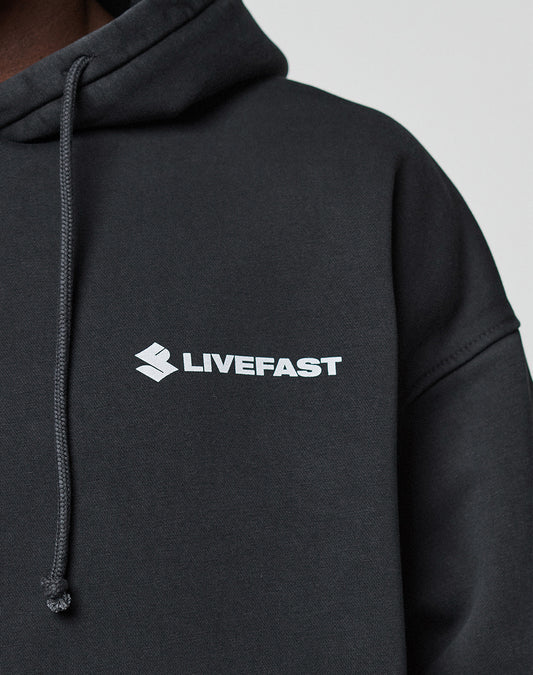A close-up shows a person in the black LFDY Dealership Hooded by LIVE FAST DIE YOUNG, made from Baumwolle, with a white LIVEFAST logo and emblem on the chest. The oversized hoodie has a drawstring and is set against a light background.