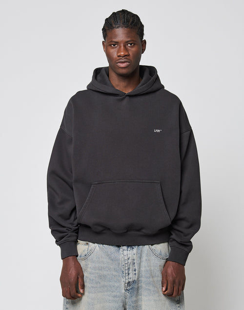 A man with braided hair wears a loose-fitting black LFDY Monaco Boys Hooded by LIVE FAST DIE YOUNG and light blue jeans, standing against a plain light gray background. The hoodie is crafted from Baumwolle Polyester for comfort.