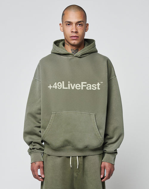 A person with a shaved head and neck tattoo wears the Basic +49 Hooded by LIVE FAST DIE YOUNG—an olive green, oversized hoodie with +49LiveFast on the front, paired with matching faded sweatpants, standing against a plain light gray background.