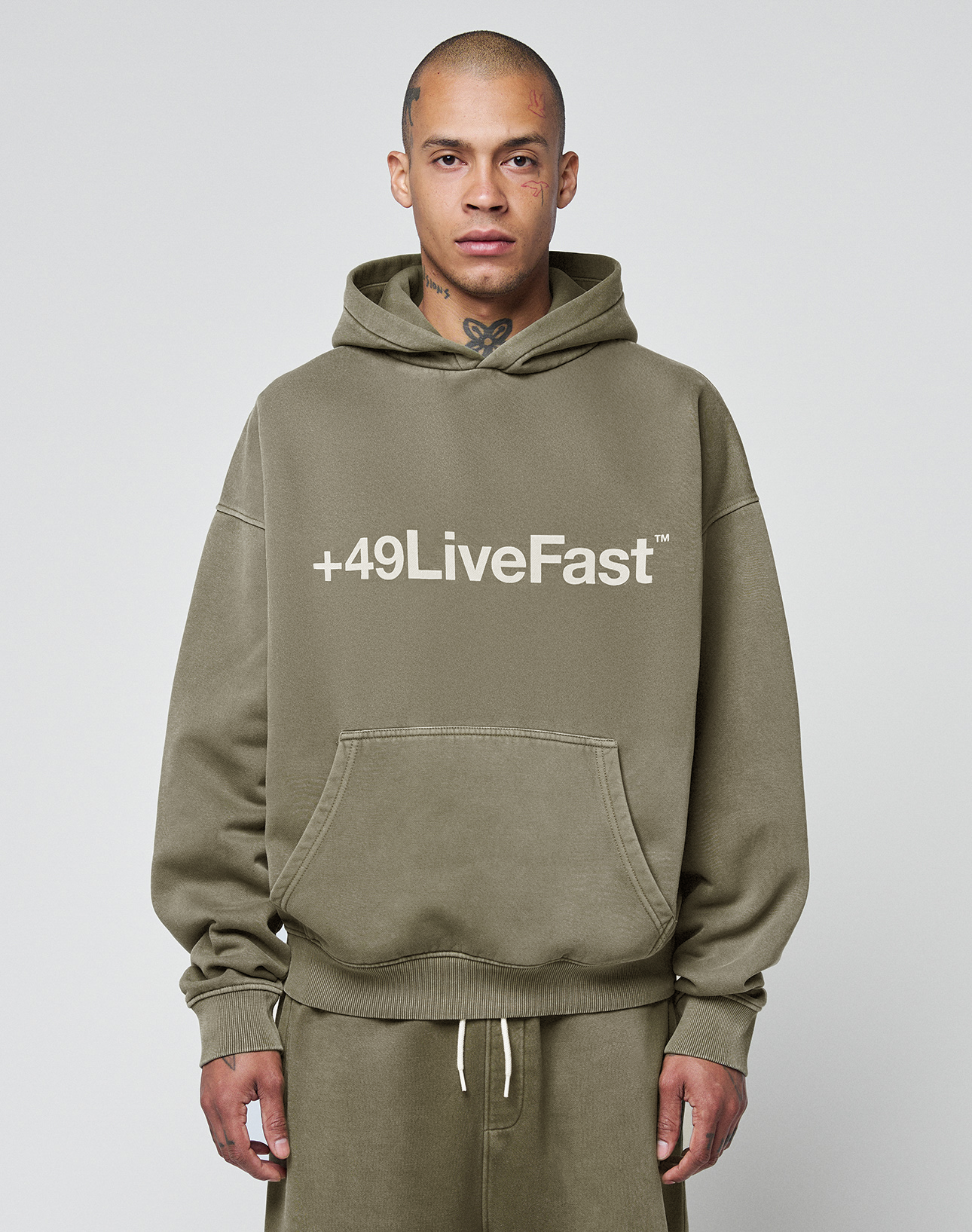 A person with a shaved head models the Basic +49 Hooded by LIVE FAST DIE YOUNG in olive green with white +49LiveFast text, matching sweatpants, and a relaxed oversize fit and faded look against a plain light gray background.
