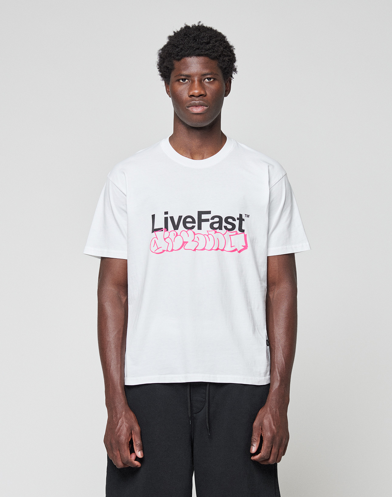 A man stands against a grey background, wearing the LIVE FAST DIE YOUNG Basic Live Fast Graffiti Tee—100% Baumwolle, white with bold "LiveFast" frontprint and pink graffiti-style text—paired with black pants for a regular fit look.