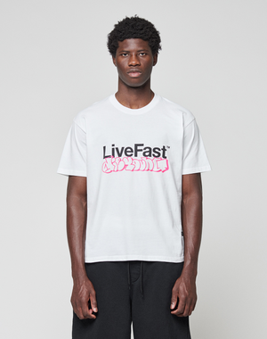 A man stands against a grey background, wearing the LIVE FAST DIE YOUNG Basic Live Fast Graffiti Tee—100% Baumwolle, white with bold "LiveFast" frontprint and pink graffiti-style text—paired with black pants for a regular fit look.