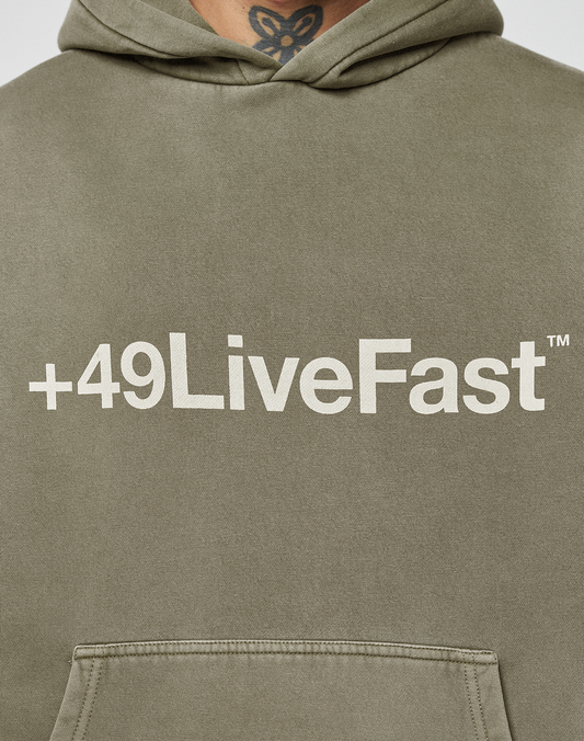 A person in the Basic +49 Hooded by LIVE FAST DIE YOUNG—a hand-faded, oversized olive green hoodie with white +49LiveFast™ text across the chest. Part of a neck tattoo appears above the hood.