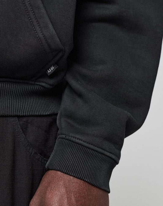 Close-up of a person in a black LFDY Dealership Hooded sweatshirt and black pants. Focus on the ribbed cuff, hand, and small LEDU label. Crafted by LIVE FAST DIE YOUNG from soft cotton for ultimate comfort.