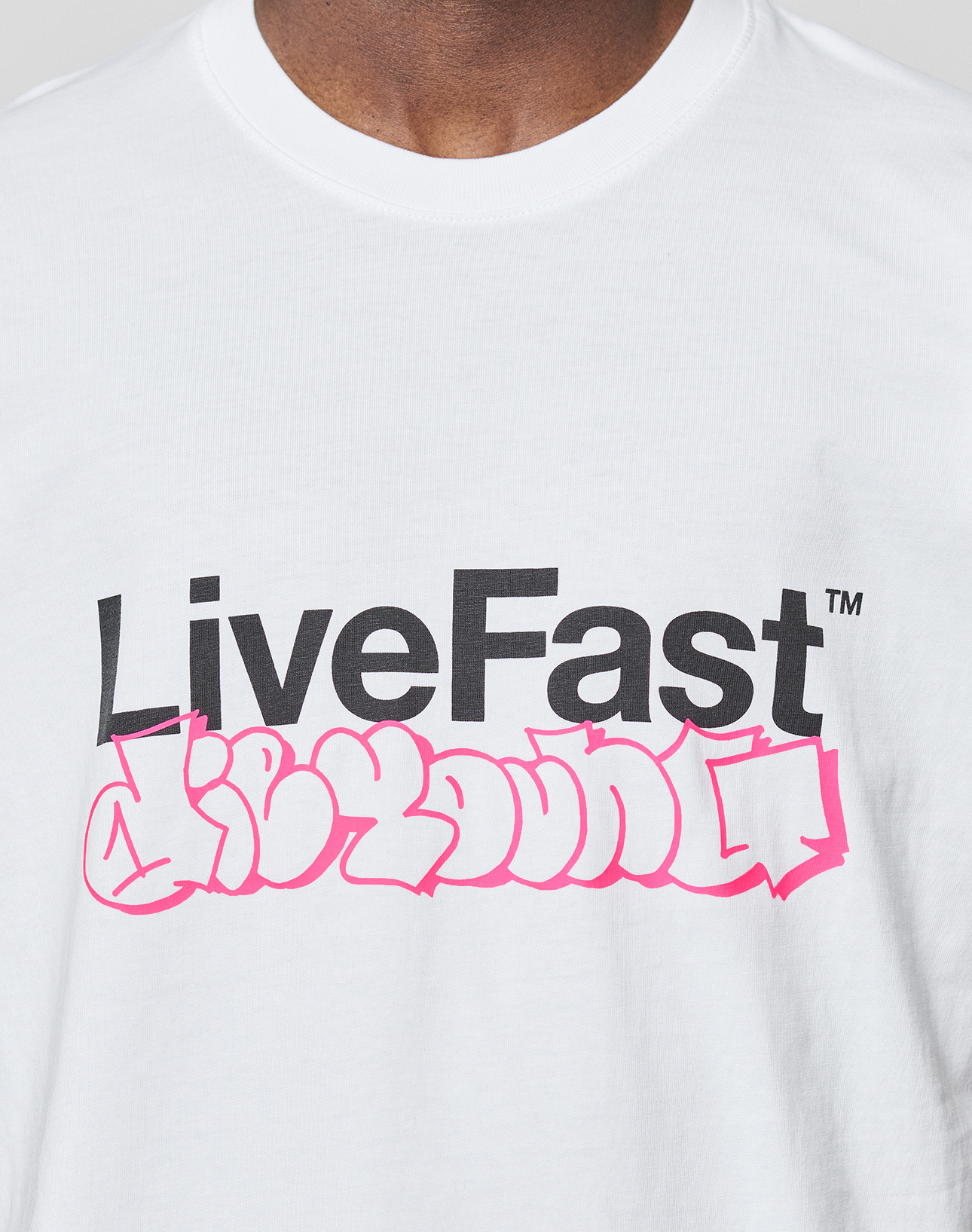 A person wearing the LIVE FAST DIE YOUNG Basic Live Fast Graffiti Tee, a regular fit white 100% Baumwolle T-shirt featuring a bold black "LiveFast" frontprint above pink graffiti-style writing.