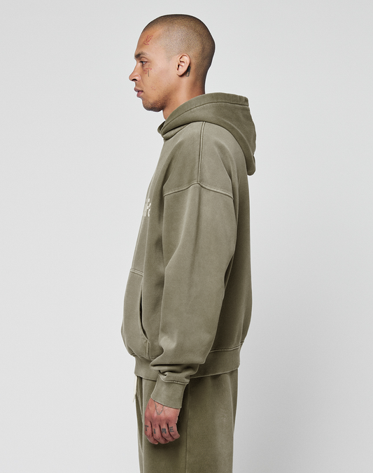A person stands in profile against a plain white background wearing the LIVE FAST DIE YOUNG Basic +49 Hooded in olive green with a handgefertigter Fade-Effekt and matching pants. The hoodie features a front pocket, hood down, and facial tattoos are visible.