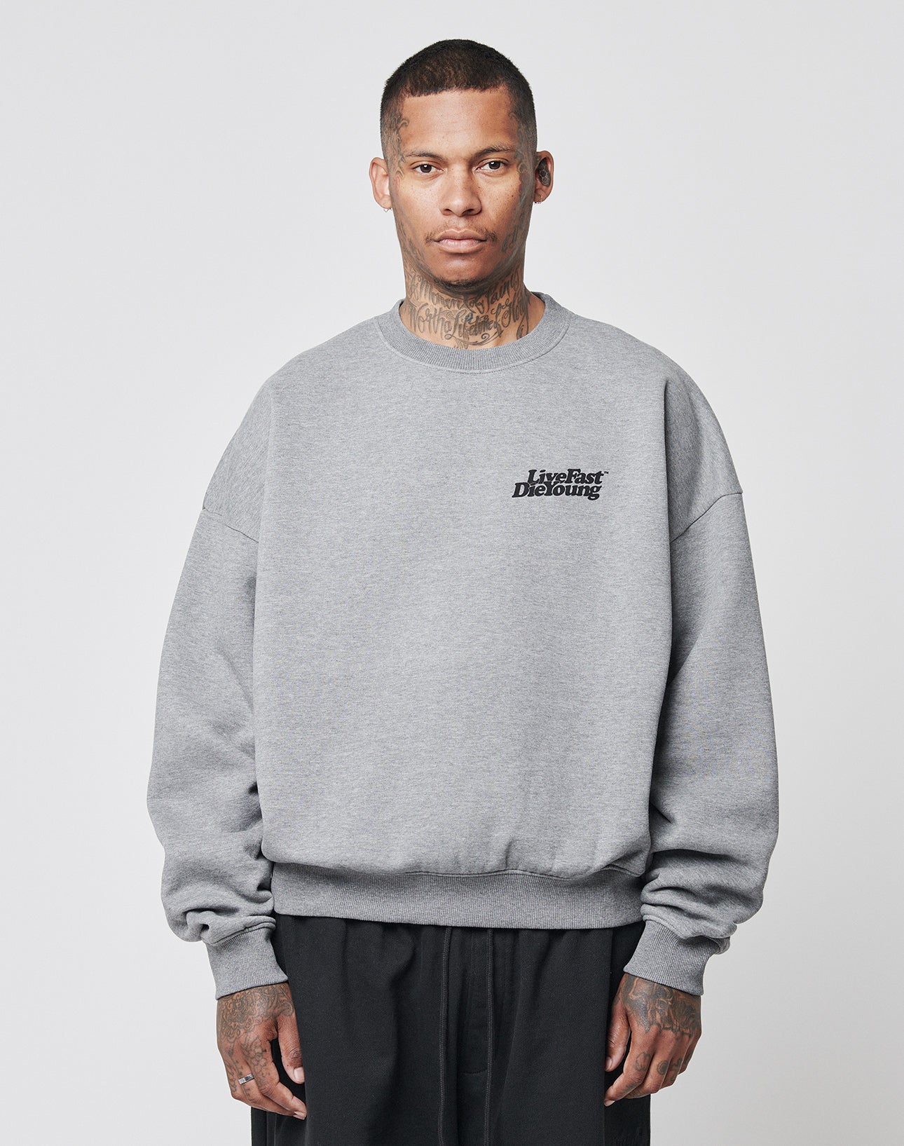 A person with short hair and visible neck tattoos wears the Basic 360 Sweater by LIVE FAST DIE YOUNG, featuring a loose fit and bold front and back prints, paired with black pants against a plain light background.