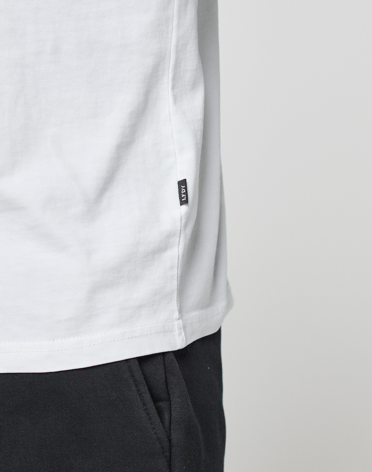 Close-up of a person wearing the LIVE FAST DIE YOUNG Basic Live Fast Graffiti Tee (100% Baumwolle, white, small black side tag) with black pants, standing against a plain light gray background.