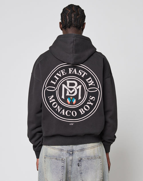 A person with braided hair, seen from behind, wears an oversize fit black LIVE FAST DIE YOUNG LFDY Monaco Boys Hooded made from Baumwolle Polyester, featuring a large circular crest backprint, paired with light blue jeans.