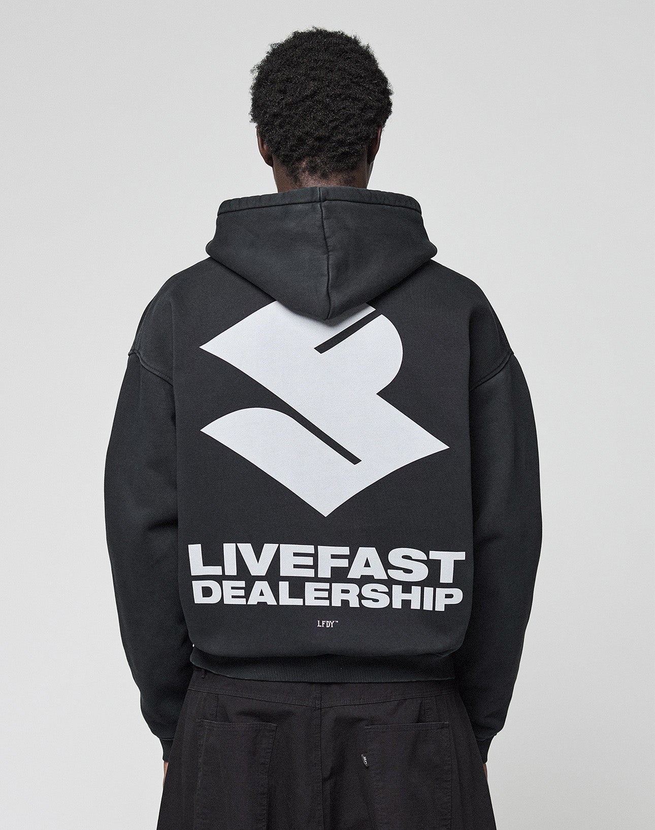 A person with short, curly hair is seen from behind in the black LFDY Dealership Hooded by LIVE FAST DIE YOUNG, featuring an oversize fit with a large white geometric logo and bold LIVEFAST DEALERSHIP text as a backprint.