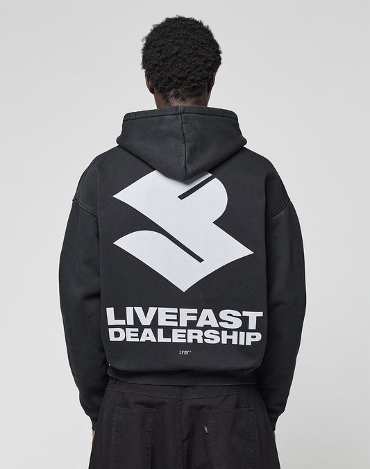 A person with short, curly hair is seen from behind in the black LFDY Dealership Hooded by LIVE FAST DIE YOUNG, featuring an oversize fit with a large white geometric logo and bold LIVEFAST DEALERSHIP text as a backprint.
