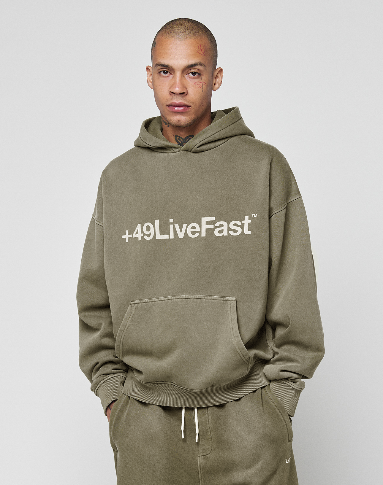 A person faces forward in an olive green Basic +49 Hooded and matching Oversize Fit sweatpants from LIVE FAST DIE YOUNG. The hoodie features a large front pocket and “‘+49LiveFast™’” in white on the chest against a plain, light background.