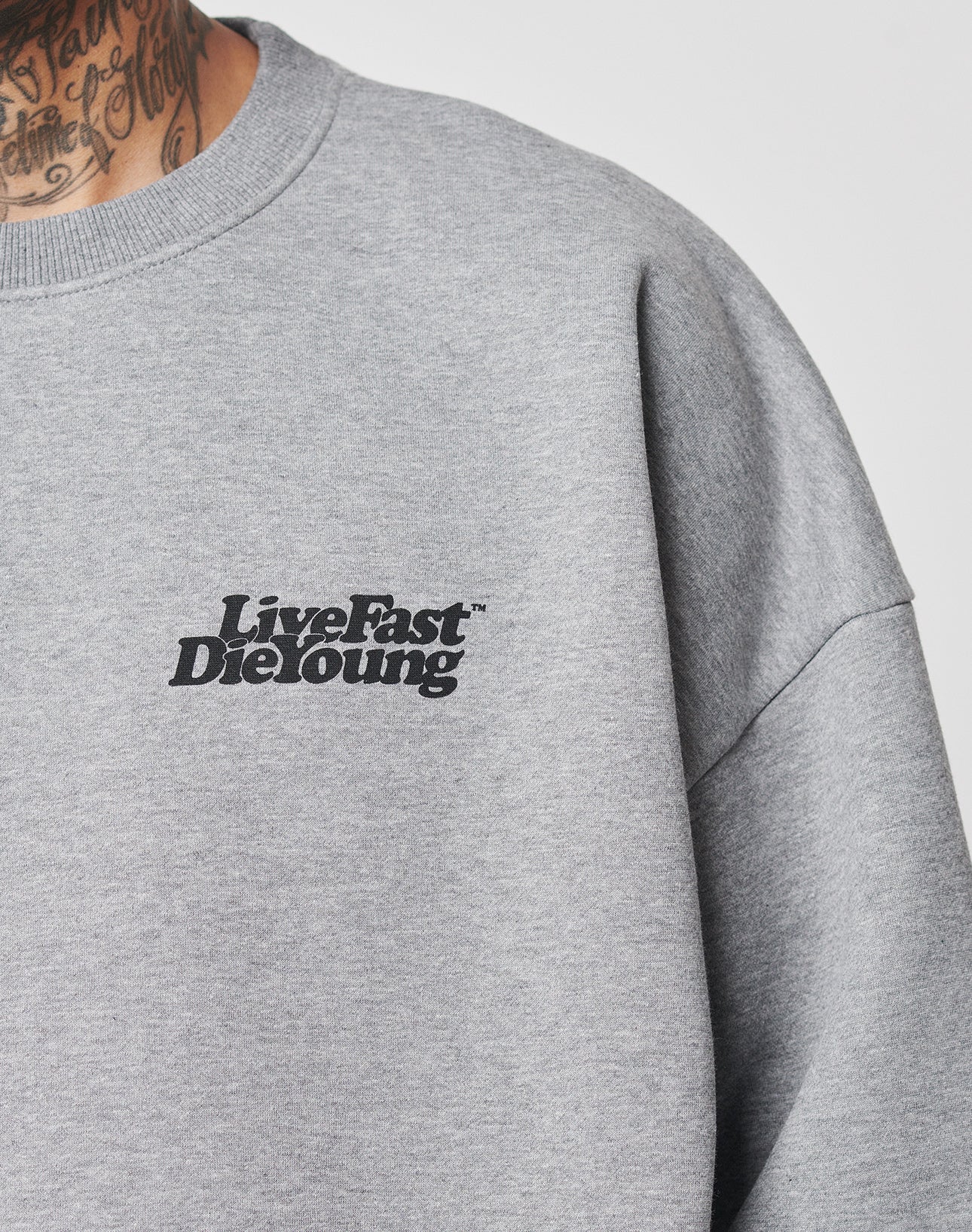 Close-up of a person wearing the Basic 360 Sweater by LIVE FAST DIE YOUNG, featuring a grey color, black Live Fast Die Young front print, oversize fit, visible topstitching, and part of their tattooed neck.