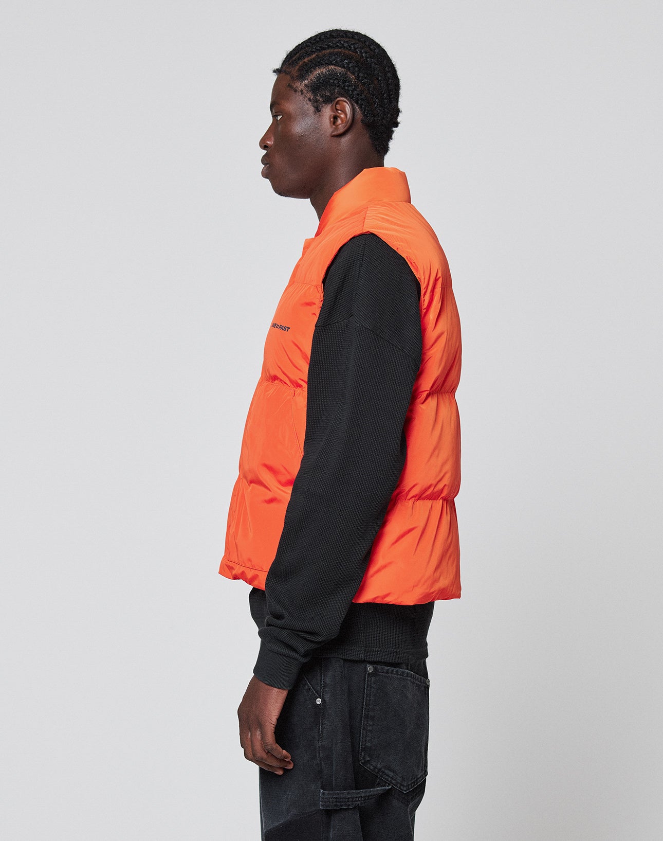 A person with braided hair stands in profile against a light gray background, wearing the LIVE FAST DIE YOUNG Live Fast Cropped Vest in bright orange over a black long-sleeve shirt and black jeans.