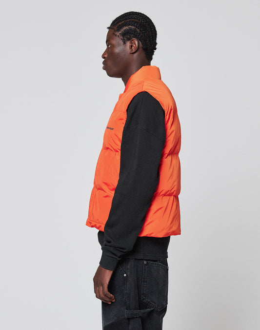 A person with braided hair stands in profile against a light gray background, wearing the LIVE FAST DIE YOUNG Live Fast Cropped Vest in bright orange over a black long-sleeve shirt and black jeans.