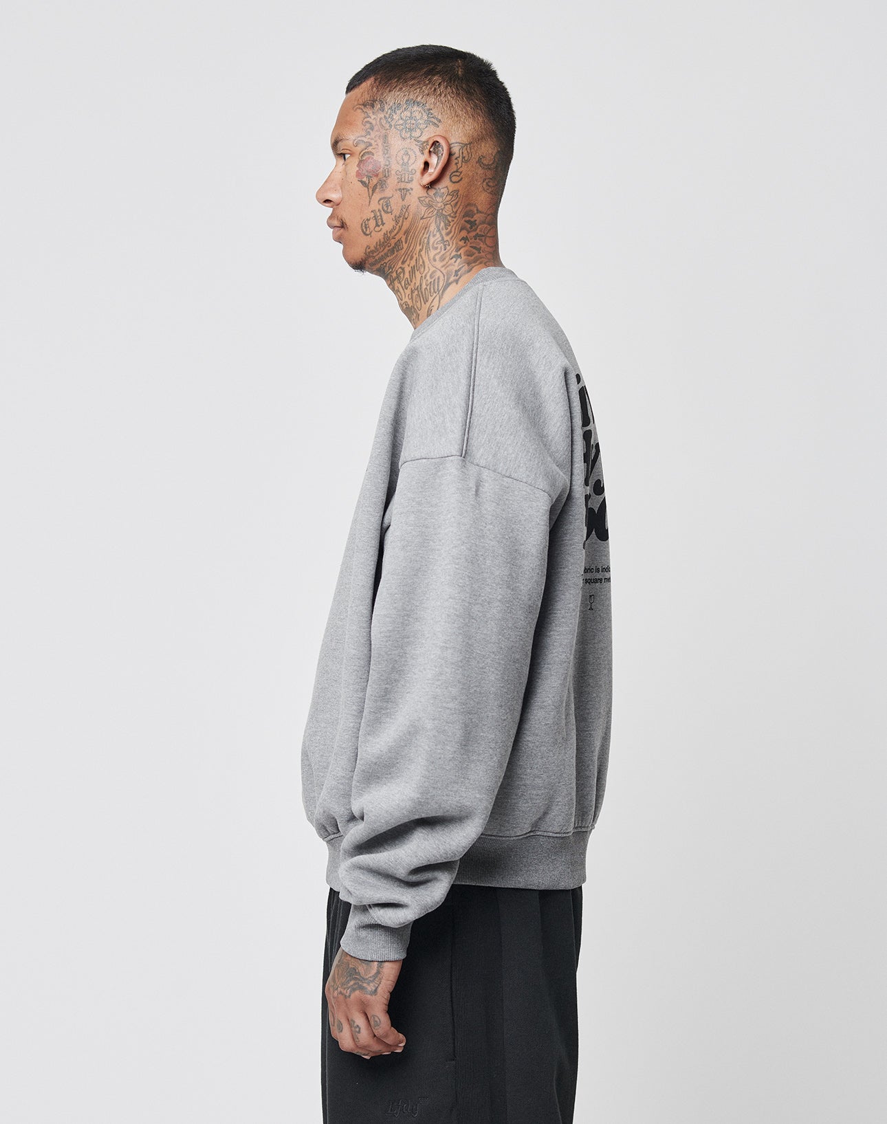 A man with extensive neck and face tattoos stands in profile, wearing the Basic 360 Sweater by LIVE FAST DIE YOUNG and black pants against a plain white background.
