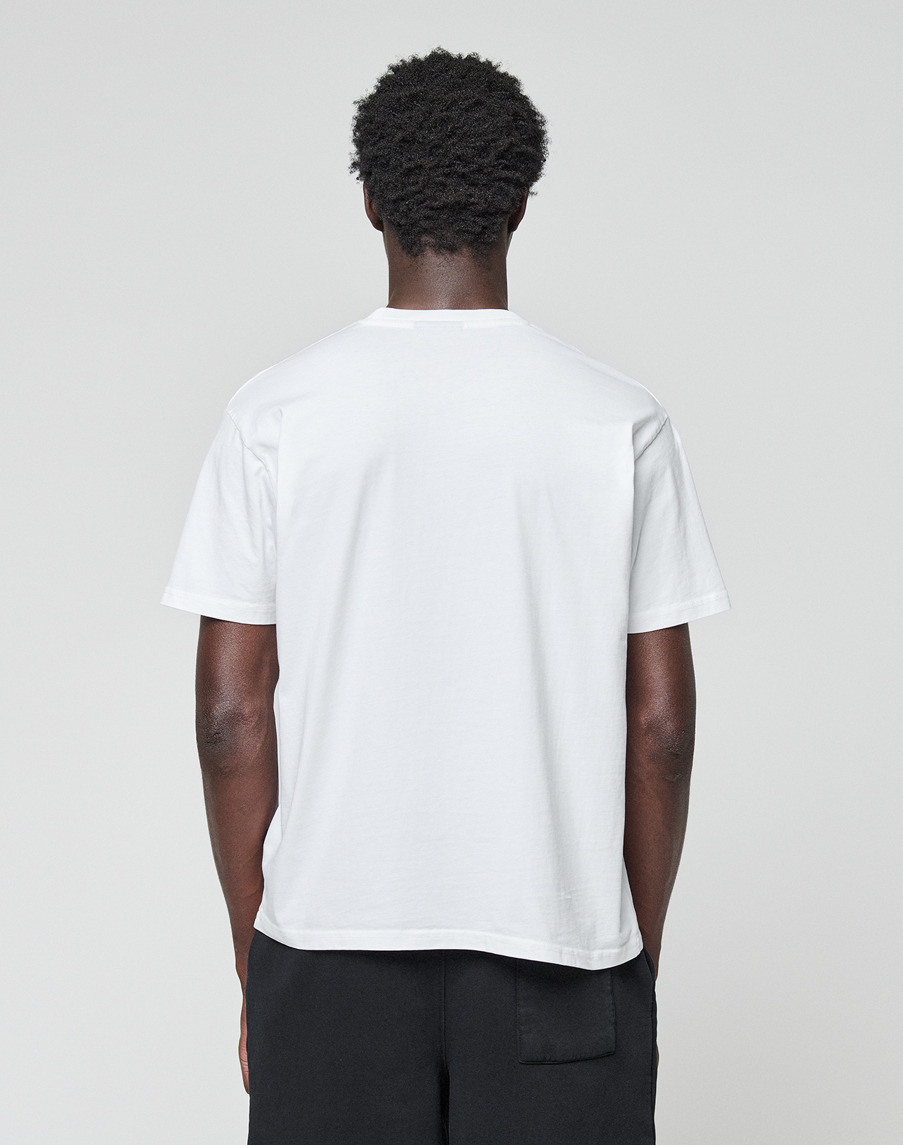 A person with short curly hair faces away from the camera, wearing the LIVE FAST DIE YOUNG Basic Live Fast Graffiti Tee and black pants against a light gray background.