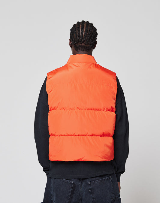 A person with braided hair, seen from behind, wears the LIVE FAST DIE YOUNG Live Fast Cropped Vest in bright orange over a black long-sleeve shirt and dark jeans, against a plain light background.