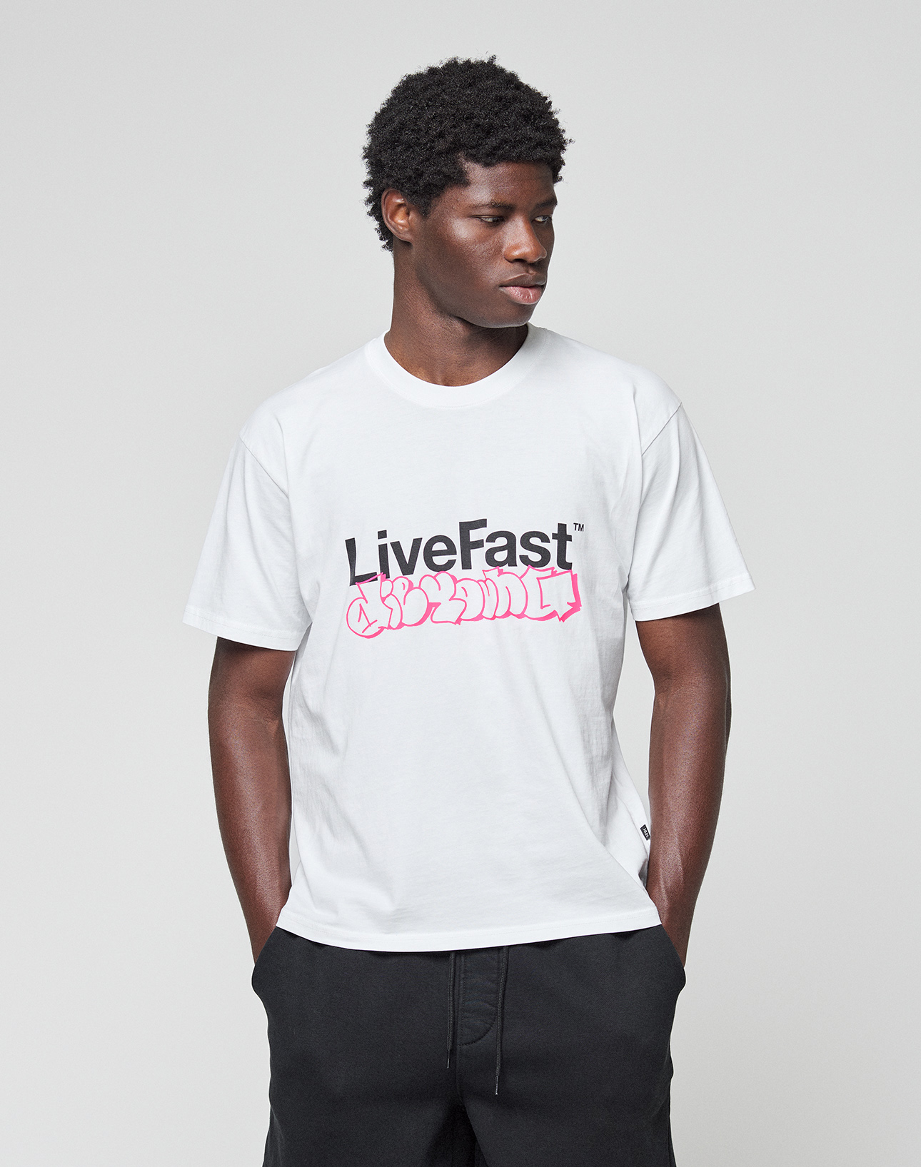 A man stands against a gray background, hands in pockets, wearing the LIVE FAST DIE YOUNG Basic Live Fast Graffiti Tee—a regular fit white T-shirt with a bold black "LiveFast" print and pink graffiti text. Made from 100% Baumwolle.
